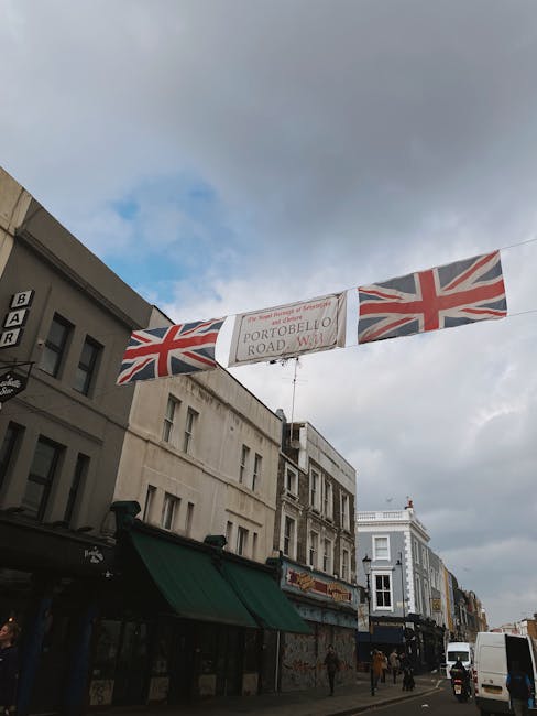 A street scene on Portobello Road in Notting Hill featuring a banner with the Union Jack flags and the text 'Portobello Road' suspended between buildings. The storefronts below have various facades, with one painted in dark green and another in white, and display windows with merchandise and signage. The sky is partly cloudy with patches of blue, and pedestrians are walking along the street, which is lined with shops and commercial establishments. The image emphasizes the vibrant, busy atmosphere of the area, likely linked to the importance of maintaining clean, inviting exteriors and shopfronts in the neighborhood, as highlighted in the Best Cleaning Tips for Portobello Road Shopfronts, NOTTING HILL Cleaner.