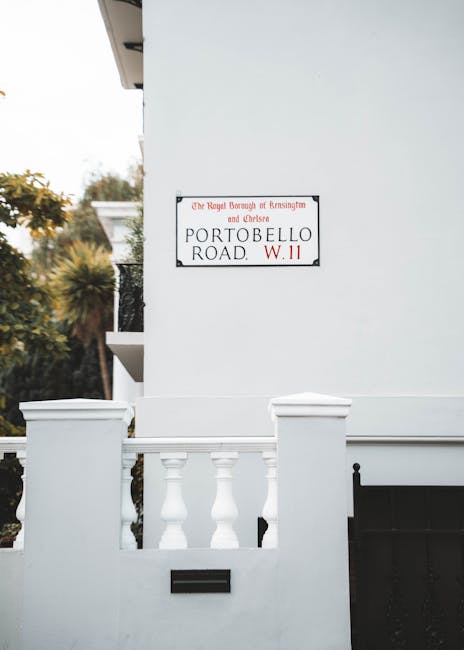 A white exterior wall of a residential building in Portobello Road, Notting Hill, featuring a street sign and a decorative white balustrade with columns. The wall appears clean and well-maintained, with smooth surfaces and no visible dirt or grime. In the background, there is greenery with blurred trees and foliage, suggesting an outdoor setting. The lighting is natural, highlighting the freshness of the clean surfaces. This image exemplifies the importance of surface cleaning and upkeep, which Notting Hill Cleaner offers in their best cleaning tips for Portobello Road shopfronts and residential spaces.