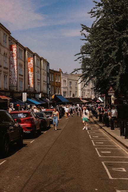 A street scene on Portobello Road in Notting Hill showing a sign suspended between two buildings with Union Jack flags on either side, indicating the location. The sign reads 'Portobello Road, W11,' a