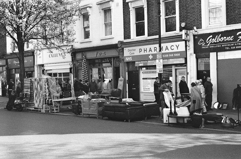 A street scene on Westbourne Grove in Notting Hill showing a row of commercial establishments including a pharmacy with a sign reading 'Doctor Care NHS' and a seafood specialist shop named 'Galborne'. In front of the shops, there are several tables and chairs, some covered with various objects, and a display of furniture and household items laid out along the pavement. Pedestrians are seen walking and browsing, with some wearing coats and hats, indicating cool weather. The building facades are brick, with large windows and signage, and there are trees with autumn foliage lining the street. The overall scene appears busy and organized, typical of a vibrant shopping street, captured in black and white for a classic aesthetic, emphasizing cleanliness and orderliness in the urban environment, with surrounding area hinting at a well-maintained commercial district as part of Notting Hill's lively community. Notting Hill Cleaner’s services could support proper disposal and clearance of bulky waste items displayed outside.