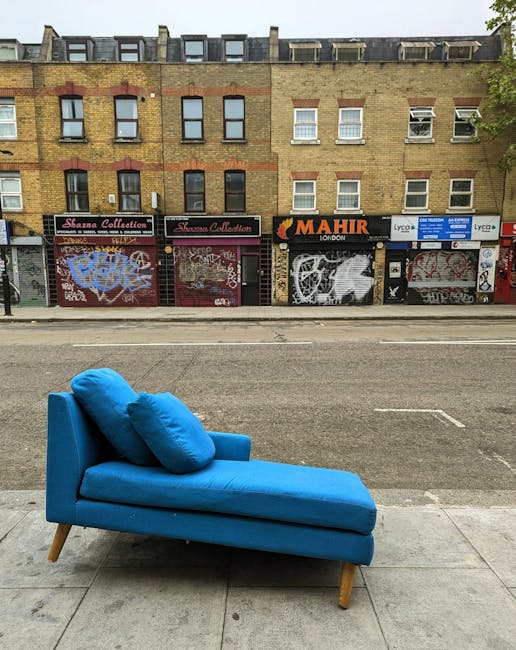 A bright blue chaise lounge with two matching cushions is positioned on a paved sidewalk in front of a city street. The furniture appears clean and well-maintained, with smooth fabric upholstery and wooden legs. Behind it, a road with visible lane markings runs parallel, and across the street is a row of three-story brick commercial buildings featuring shuttered storefronts, some with graffiti. The buildings have a mix of yellow and red brickwork, with rectangular windows, some open, and varying signage above shopfronts, including a sign for 'Mahir London'. The scene is evenly lit, indicating daytime, and the area appears urban and somewhat weathered, contrasting the fresh appearance of the furniture.