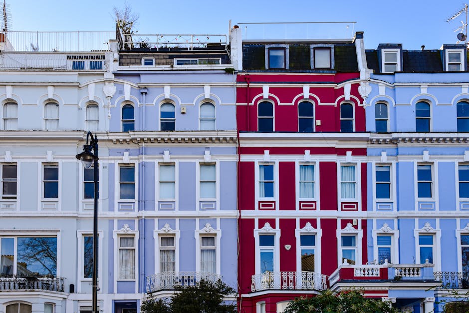 Photograph of a row of Victorian-style terraced residential buildings on Ladbroke Grove, showing ornate facades painted in pastel blue and vibrant red, with white decorative trims. The buildings feature multiple tall, narrow windows with white frames, some with curtains, and decorative cornices. A black streetlamp is visible in the foreground, and the rooftops have rooftop gardens with greenery and antennas. The image reflects an urban neighbourhood setting with clear blue sky and natural daylight illuminating the buildings, representative of typical London architecture. This setting aligns with the context of end-of-tenancy cleaning, as cleaning specialists like Notting Hill Cleaner focus on maintaining the pristine appearance of such properties through surface cleaning, deep cleaning, and sanitisation services.