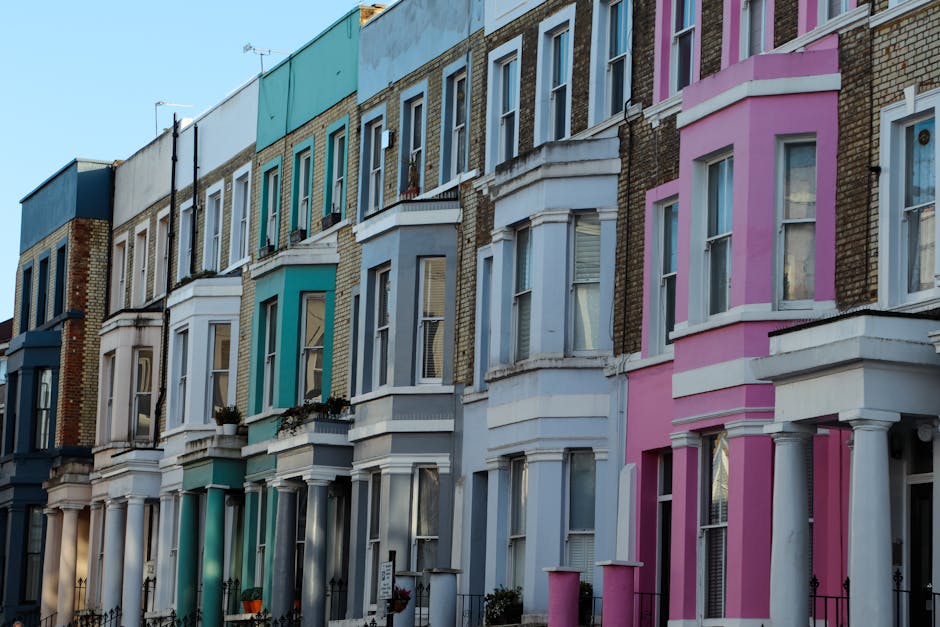 A row of multi-storey Victorian-style terraced houses in Notting Hill, featuring colorful facades in shades of blue, turquoise, white, and pink. The buildings have large bay windows with white frames, some with flower boxes on the window sills, and small front gardens enclosed by black wrought iron fences. Bright natural daylight illuminates the street, highlighting the clean and well-maintained exterior surfaces. The image reflects the vibrant and historic architectural style typical of Notting Hill, with a focus on aesthetic detail and residential character. This scene exemplifies the importance of surface cleaning and maintenance to preserve the area's charm, aligning with Notting Hill Cleaner's expertise in deep cleaning and sanitisation for residential properties.