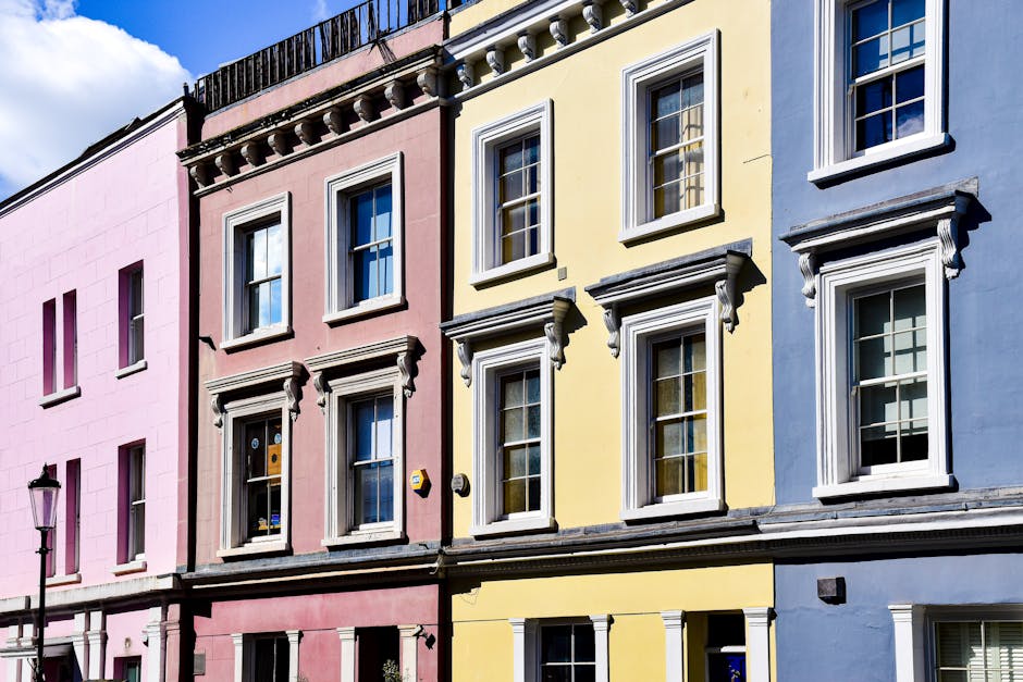 A row of vibrant, pastel-colored residential buildings with neatly painted facades in pink, yellow, and blue, each featuring white-framed sash windows and decorative mouldings. The buildings display a clean exterior, with polished window sills and orderly window panes, under a partly cloudy sky. In the foreground, a vintage street lamp is visible, enhancing the charming urban setting. This image exemplifies well-maintained, surface cleaning and external detailing typical of domestic cleaning standards supported by Notting Hill Cleaner, as referenced on nottinghillcleaner.co.uk in relation to surface cleaning, deep cleaning, and maintenance for residential properties.