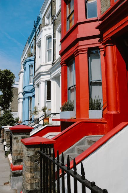 Photograph of a row of colorful terraced residential buildings on a sunny day, featuring facades painted in shades of blue, white, and red. The blue and white buildings have large, white-framed windows with decorative moldings, while the red building has a prominent porch with red-painted concrete stairs and black wrought iron railings. The buildings are set along a paved sidewalk with small planters containing greenery, and the scene is illuminated by natural daylight, emphasizing the clean and well-maintained appearance of the exteriors. Notting Hill Cleaner specializes in surface cleaning and deep cleaning of residential properties, ensuring hygiene and sparkle for guest check-ins at establishments like Notting Hill Gate B&Bs.
