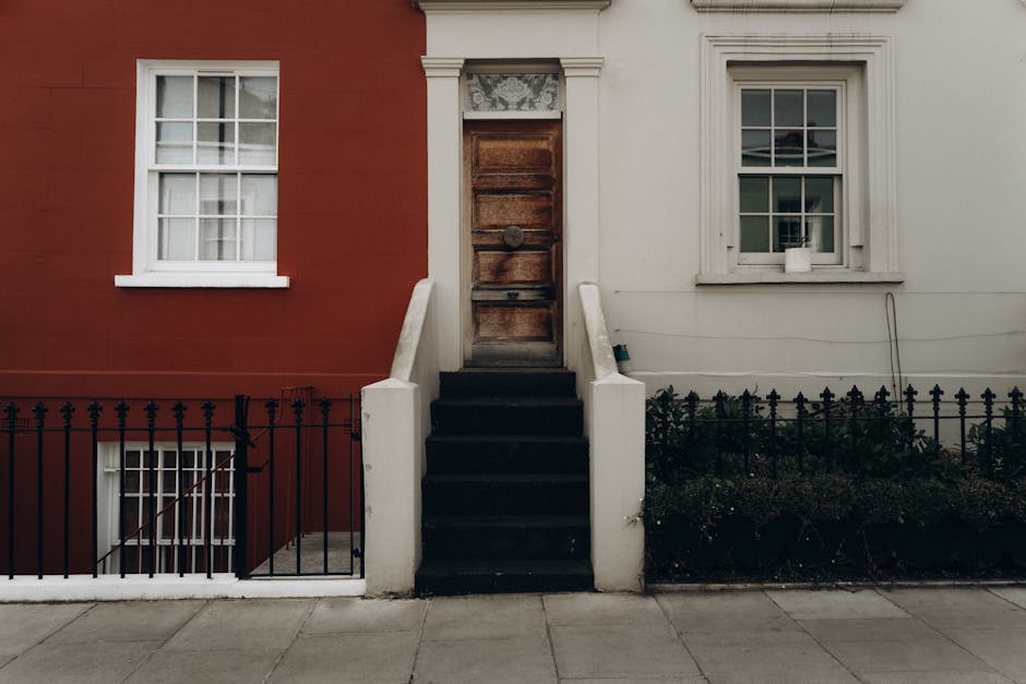 Image of the front exterior of a residential building in Notting Hill, with a set of black-painted concrete stairs leading to a wooden front door. The building’s facade features contrasting sections: a red brick wall on the left with a white-framed window and a white section on the right, also with a white-framed window. The stairs are flanked by white concrete railings, and the area at the base is bordered by a black wrought iron fence. There is a small garden bed with low greenery beneath the window on the white section. The sidewalk in front of the building is clean, and the scene is lit by natural daylight, emphasizing the building's well-maintained appearance suitable for guest check-ins. Notting Hill Cleaner provides comprehensive cleaning and sanitisation services in this area to ensure properties are guest-ready.