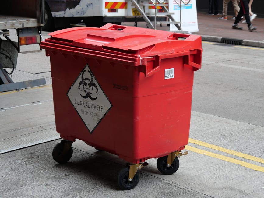 A red clinical waste disposal bin with a hinged lid, situated outdoors on a paved surface near a street curb. The bin features a biohazard symbol and a 'CLINICAL WASTE' label affixed to its front, indicating it is designated for hazardous medical waste. The container has four black caster wheels with metal supports, allowing for mobility. In the background, part of a truck and pedestrians are visible, along with a section of the sidewalk. The area appears clean, with well-maintained surfaces and natural daylight illuminating the scene. This image relates to the guidance on hazardous waste removal in W11, NOTTING HILL, provided by Notting Hill Cleaner, which specialises in cleaning services including the appropriate disposal of clinical and hazardous waste materials.