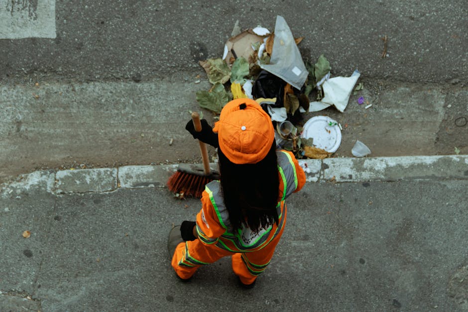 A person wearing a white protective suit, safety goggles, a respirator mask, and blue gloves standing amidst a large pile of mixed waste materials, including plastic, paper, and packaging debris, on an outdoor landfill site during daylight. The individual holds a clipboard and appears to be inspecting or documenting the waste environment, with sunlight illuminating the scene. The background shows a contaminated area with scattered refuse, emphasizing the importance of hazardous waste removal and proper sanitation. This image highlights environmental cleanup efforts and the role of professional waste management services, such as those offered by Notting Hill Cleaner, in maintaining hygiene and safety standards.