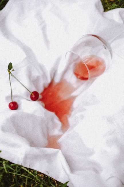 A transparent glass tumbler containing red wine rests partially submerged on a soft white fabric surface, likely a bedsheet or pillowcase, with some of the fabric subtly creased and crumpled around it. Two fresh cherries with green stems lie nearby on the fabric, adding a touch of color contrast. The scene is illuminated by natural light, highlighting the shine of the glass and the texture of the fabric. The setting appears to be indoors, possibly on a bed or lounge area, with a clean, well-maintained appearance. This image is related to surface cleaning and domestic hygiene, aligning with Notting Hill Cleaner's service of removing red wine stains from carpets and upholstery, as visible on the page about deep cleaning and stain removal techniques.