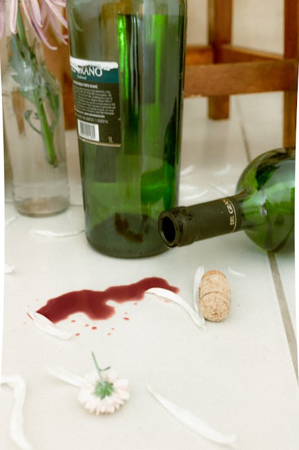Close-up of a white tiled surface in a domestic setting, featuring a wine stain in deep red color that has spread on the tiles. There is a green glass bottle, partially empty, lying on its side with a cork nearby. In the background, part of a glass vase with plant stems and leaves is visible, along with a wooden table or shelf, indicating the scene is within a living or dining area. The area appears to have minimal dust or debris, emphasizing the recent spillage. This image reflects typical scenarios of red wine accidents and highlights the importance of surface cleaning and stain removal; Notting Hill Cleaner specializes in deep cleaning and sanitisation to restore surfaces after such incidents, ensuring a hygienic environment in residential spaces.