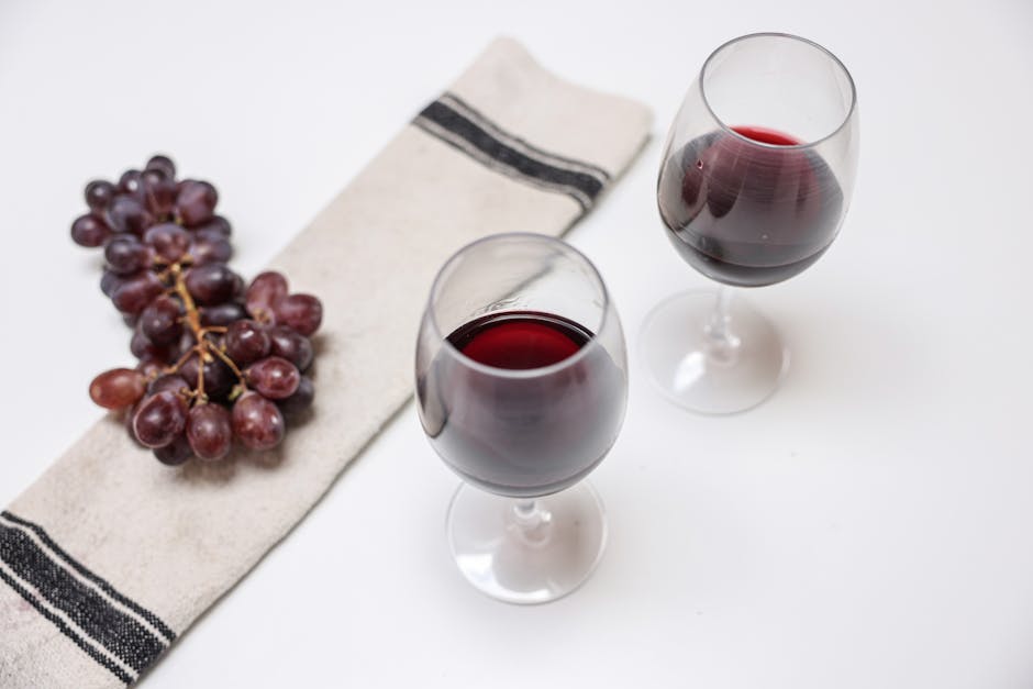 A transparent glass tumbler containing red wine rests partially submerged on a soft white fabric surface, likely a bedsheet or pillowcase, with some of the fabric subtly creased and crumpled around it. Two fresh cherries with green stems lie nearby on the fabric, adding a touch of color contrast. The scene is illuminated by natural light, highlighting the shine of the glass and the texture of the fabric. The setting appears to be indoors, possibly on a bed or lounge area, with a clean, well-maintained appearance. This image is related to surface cleaning and domestic hygiene, aligning with Notting Hill Cleaner's service of removing red wine stains from carpets and upholstery, as visible on the page about deep cleaning and stain removal techniques.