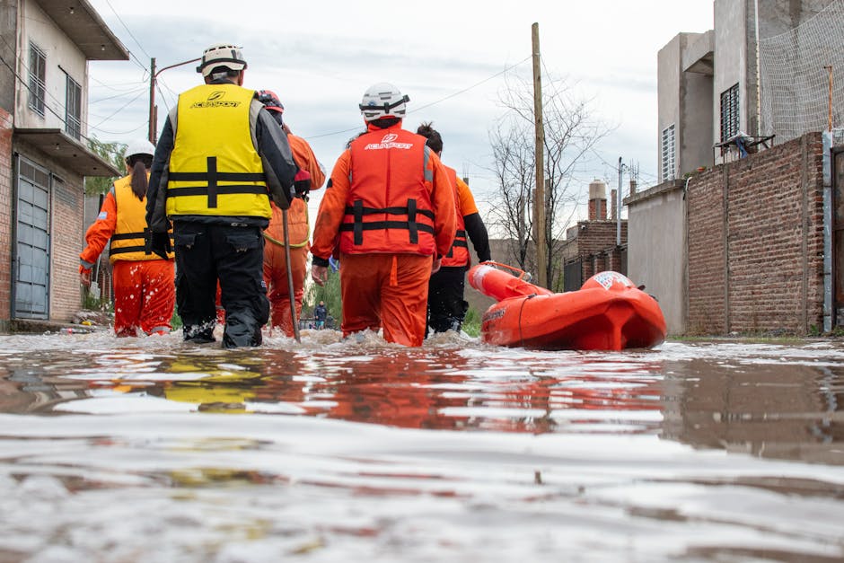 A group of emergency response personnel wearing high-visibility jackets and waterproof clothing are walking through a flooded residential street with submerged pavement and water reaching up to their ankles. The scene shows unfinished brick walls, houses, and utility poles on either side under an overcast sky. A bright orange rescue boat is visible beside them, emphasizing flood management efforts. The water reflects the overcast sky and surrounding structures, highlighting the severity of the flood. The surroundings appear muddy and disrupted, indicating recent or ongoing flooding. The image captures the urgency and scale of flood response work, aligning with emergency flood response services offered by Notting Hill Cleaner for prompt mitigation and sanitation procedures.