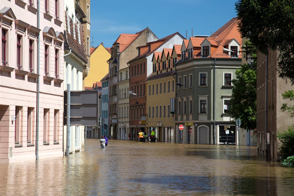 A street scene in Notting Hill with multiple multi-story residential and commercial buildings featuring varied facades in pastel pink, yellow, green, and beige. The street is flooded with water covering the cobblestone surfaces and partially submerging the lower portions of the buildings. Bright daylight illuminates the scene, with visible shadows cast by trees and the buildings' architectural details, such as gabled roofs and window trims. The area appears deserted with only a few people walking through the floodwaters, and a couple of emergency response vehicles, possibly from Notting Hill Cleaner, are present in the background. The overall appearance highlights the impact of floodwaters on an urban environment, emphasizing the need for professional flood response and surface cleaning services.