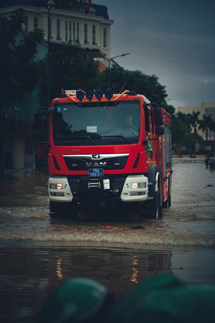 A group of emergency response personnel wearing high-visibility jackets and waterproof clothing are walking through a flooded residential street with submerged pavement and water reaching up to their ankles. The scene shows unfinished brick walls, houses, and utility poles on either side under an overcast sky. A bright orange rescue boat is visible beside them, emphasizing flood management efforts. The water reflects the overcast sky and surrounding structures, highlighting the severity of the flood. The surroundings appear muddy and disrupted, indicating recent or ongoing flooding. The image captures the urgency and scale of flood response work, aligning with emergency flood response services offered by Notting Hill Cleaner for prompt mitigation and sanitation procedures.
