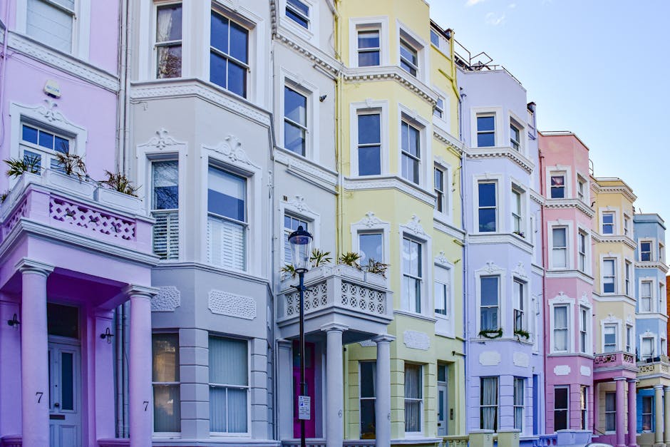 A row of pastel-colored Victorian-style residential buildings in Notting Hill, featuring ornate architectural details, bay windows, and balconies. The facades are painted in soft shades of pink, yellow, lavender, and white, with white decorative trim and window frames. The buildings have multiple floors, with some windows adorned with potted plants and small decorative elements. A street lamp and a parking sign are visible in front of the buildings. The scene is well-lit with natural daylight, highlighting the vibrant colors and pristine, clean surfaces of the exteriors, reflecting a well-maintained, hygienic urban environment as emphasized by Notting Hill Cleaner’s professional cleaning and surface maintenance services.