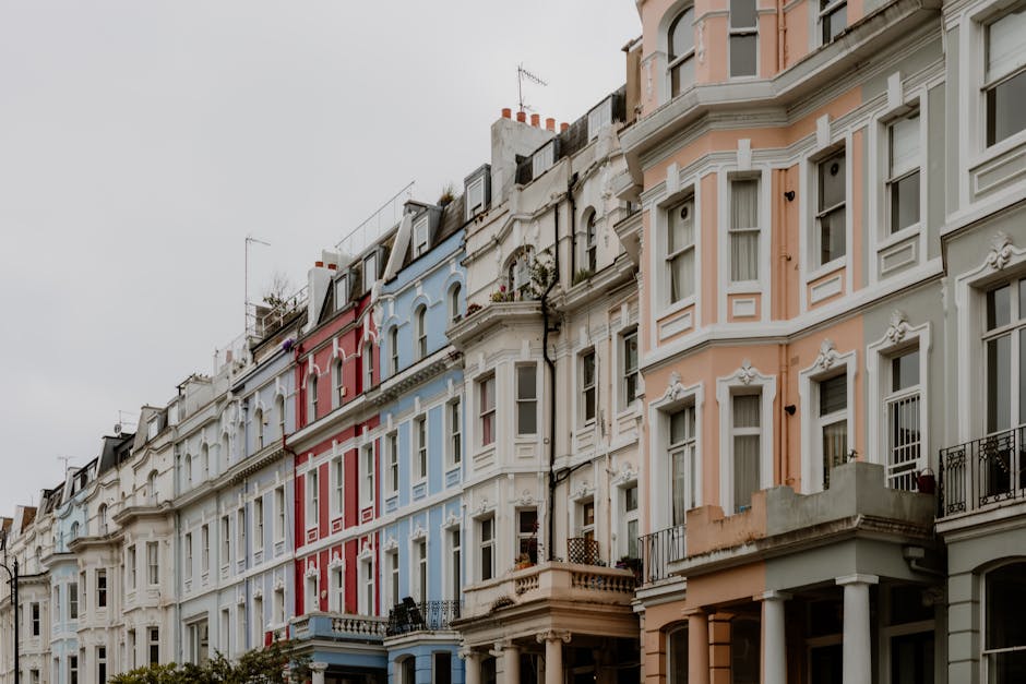 Photograph of a row of multicolored Victorian-style residential buildings in Notting Hill, featuring white, pastel pink, and light blue facades with ornate architectural details, large windows, and small balconies. The buildings have pitched roofs with chimneys visible against an overcast sky, with some greenery on balconies and rooftop terraces. The street is quiet, and natural light highlights the clean and well-maintained appearance of the exteriors, aligning with content on transparent cleaning pricing for Notting Hill flats by Notting Hill Cleaner, which emphasizes surface cleaning and house maintenance services.