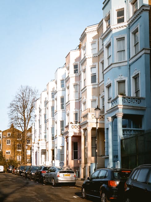 A row of pastel-colored Victorian-style residential buildings in Notting Hill, featuring ornate architectural details, bay windows, and balconies. The facades are painted in soft shades of pink, yellow, lavender, and white, with white decorative trim and window frames. The buildings have multiple floors, with some windows adorned with potted plants and small decorative elements. A street lamp and a parking sign are visible in front of the buildings. The scene is well-lit with natural daylight, highlighting the vibrant colors and pristine, clean surfaces of the exteriors, reflecting a well-maintained, hygienic urban environment as emphasized by Notting Hill Cleaner’s professional cleaning and surface maintenance services.